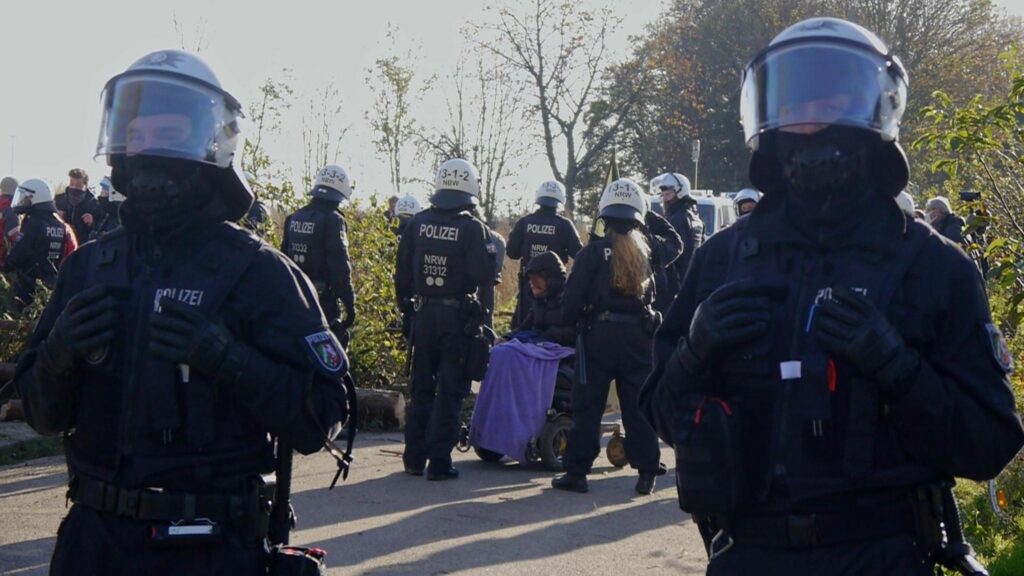 Eine Person im E-Rollstuhl auf einer Straße umringt von vielen Polizist*innen in Riot-Ausrüstung.