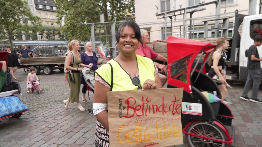 Sana, Eelam Tamil Aktivisti mit schwarzen Haaren, Ordnerweste und Schild: "Behinderte Geflüchtete, leave no-one behind"