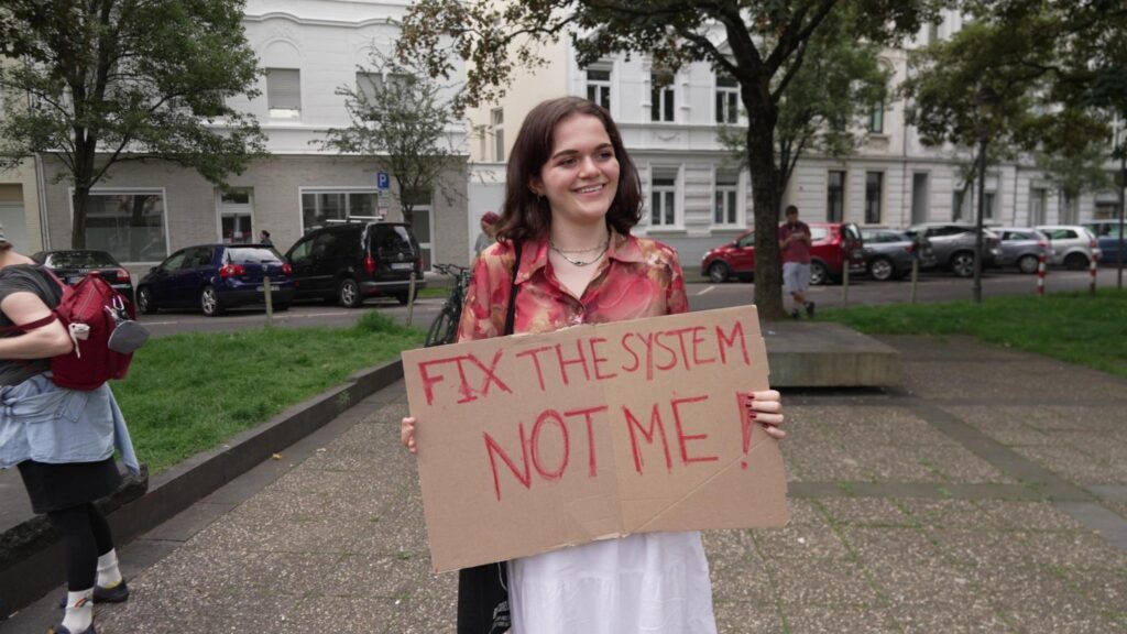 Person mit langen braunen Haaren und weißem Rock hält Schild "Fix the system not me!"