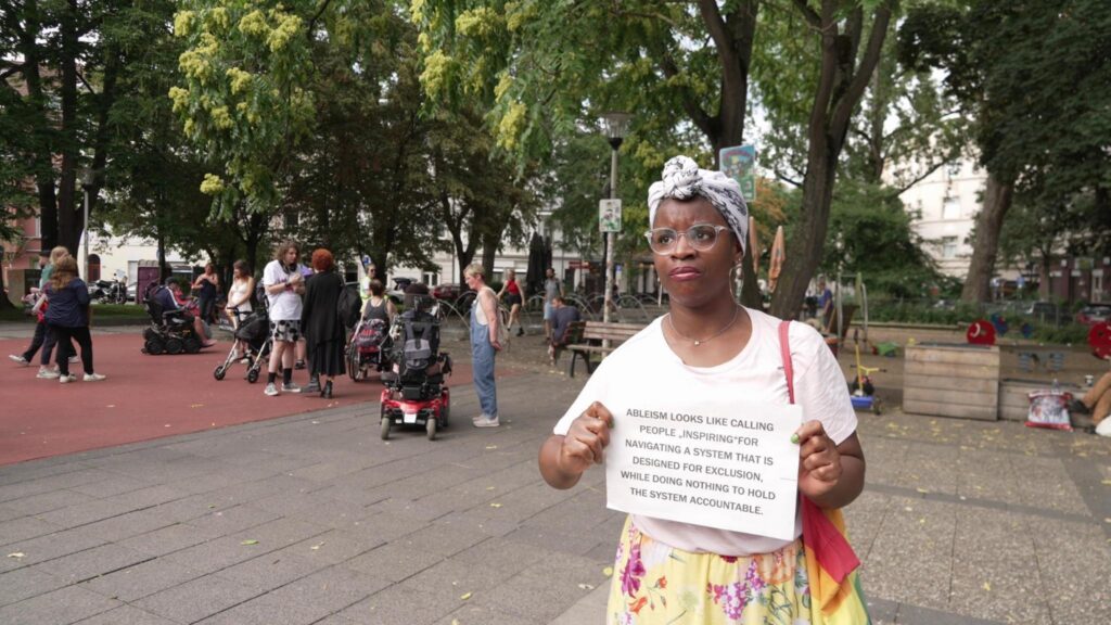 Tabitha, bunt gekleidete Schwarze Künstler*in mit Regenbogentasche und weissem vorne geknotetem Kopftuch hält einen Schild "Ableism looks like calling people 'inspiring' for navigating a system that is designed for exclusion, while doing nothing to hold the system accountable".