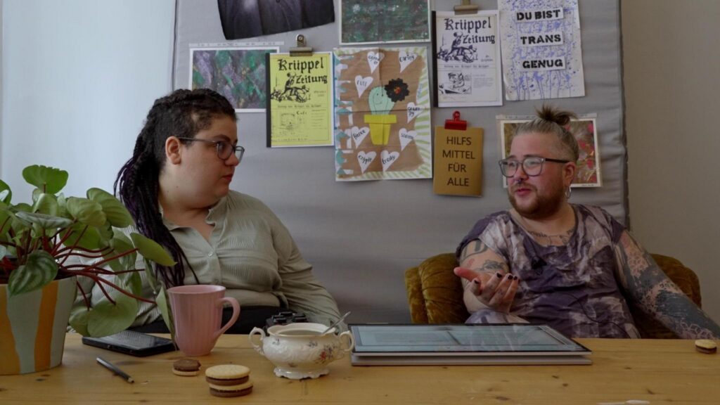 SchwarzRund und simo tier sitzen im Atelier am Tisch und reden. Dahinter Poster und Zines auf die Wand, darunter: "Hilfsmittel für Alle", "Du bist trans genug", "Krüppel-Zeitung"