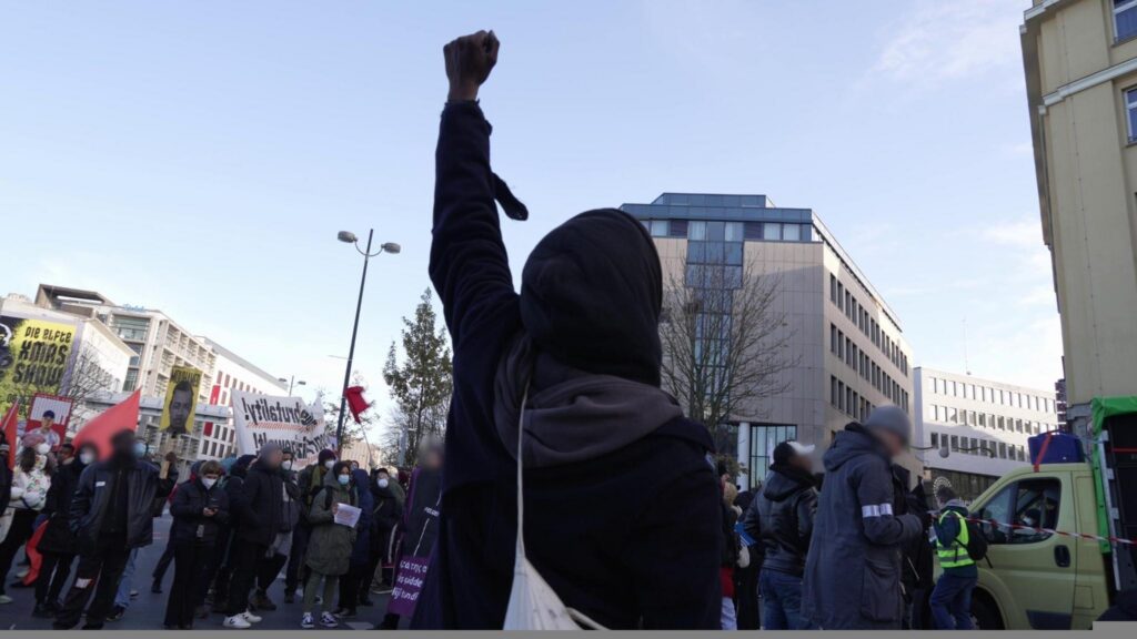 Ein Mensch in einer Demonstration hält die linke Faust hoch in den Himmel. Dahinter ein Lautsprecherwagen und viele Menschen mit Banner und Schilder, "Justice for Mouhamed" ist lesbar.